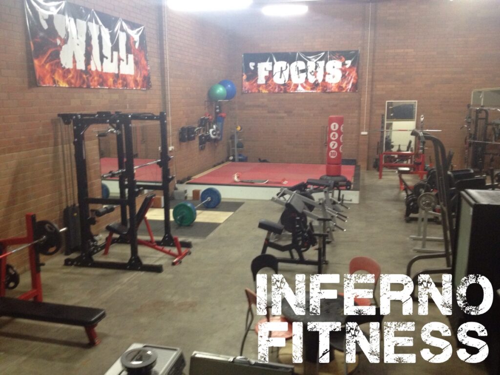 A gym with various weightlifting equipment, benches, and platforms. Banners on the wall read WILL and FOCUS. The words INFERNO FITNESS are prominently displayed in the foreground.
