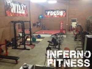 A gym with various weightlifting equipment, benches, and platforms. Banners on the wall read WILL and FOCUS. The words INFERNO FITNESS are prominently displayed in the foreground.