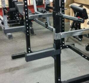 A gym setup featuring weightlifting equipment, including a squat rack with adjustable safety bars and a bench in the background. The floor is covered with dark mats and various weights are visible.