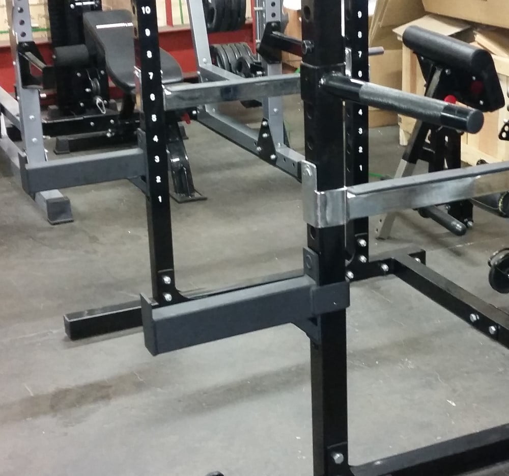 A gym setup featuring weightlifting equipment, including a squat rack with adjustable safety bars and a bench in the background. The floor is covered with dark mats and various weights are visible.