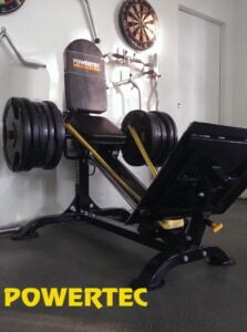 A leg press machine with multiple weight plates is positioned against a wall. A dartboard hangs on the wall above the machine, and a yellow POWERTEC logo is visible on the machine and floor. Several gym hooks are mounted on the wall.