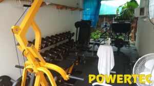 Home gym with various exercise equipment, including a yellow Powertec machine, a rack of dumbbells, a leg press, and benches. Theres a white towel on one bench and a fan nearby. The room is enclosed with natural light coming in.