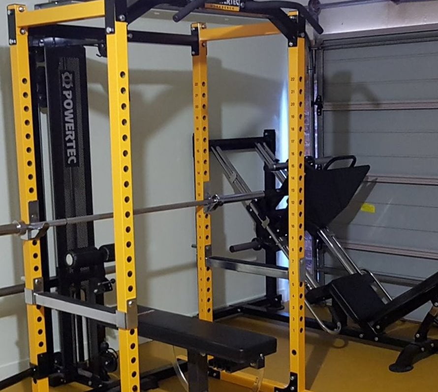 A yellow power rack equipped with a bench, barbell, and pulley system in a gym setting. Theres also a leg press machine visible in the background. The gym has a concrete wall and a partially open garage door.