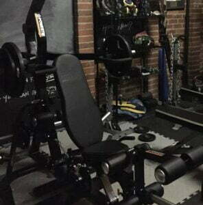 A home gym with various equipment, including a weight bench with attached weights, resistance bands, a punching bag, and mats. The room has brick walls and shelves filled with workout gear.