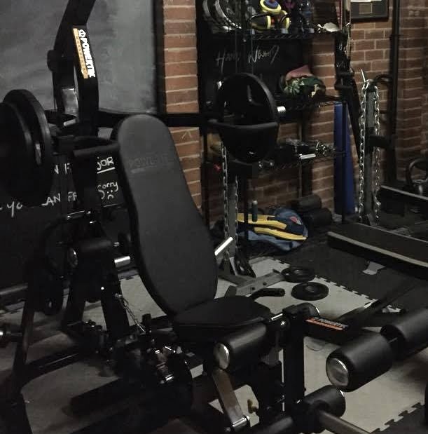 A home gym with various equipment, including a weight bench with attached weights, resistance bands, a punching bag, and mats. The room has brick walls and shelves filled with workout gear.