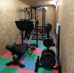 Home gym featuring a leg press machine and various weights on a rack. The room has wooden walls and a colorful foam mat flooring in red, green, and blue squares.