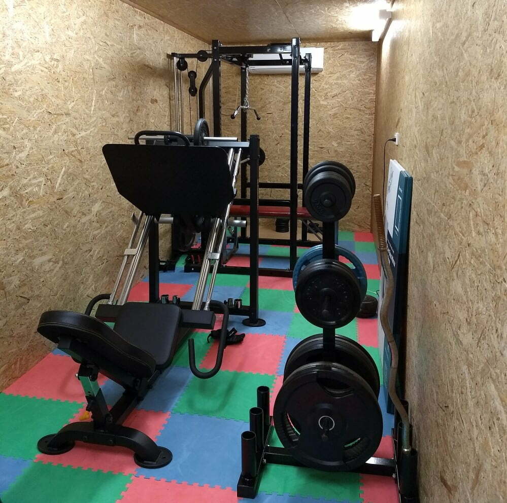 Home gym featuring a leg press machine and various weights on a rack. The room has wooden walls and a colorful foam mat flooring in red, green, and blue squares.