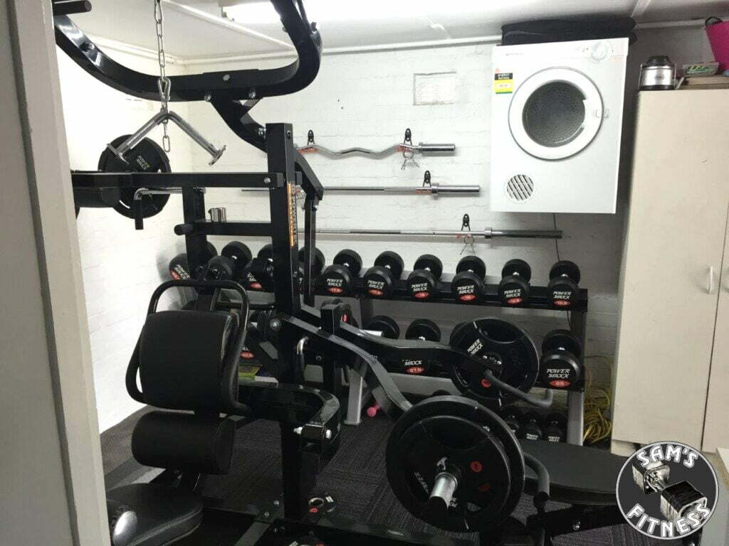 A home gym with weightlifting equipment, including a multi-functional weight machine, dumbbells on racks, and a wall-mounted dryer. The Sams Fitness logo appears in the bottom right corner.