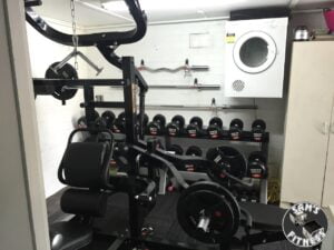 A home gym with weightlifting equipment, including a multi-functional weight machine, dumbbells on racks, and a wall-mounted dryer. The Sams Fitness logo appears in the bottom right corner.