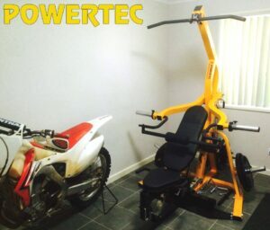 A compact room with a Powertec weight machine and a white and red dirt bike parked next to it. The weight machine is bright yellow with black padding and adjustable weights. The room has tiled flooring and a window with vertical blinds.