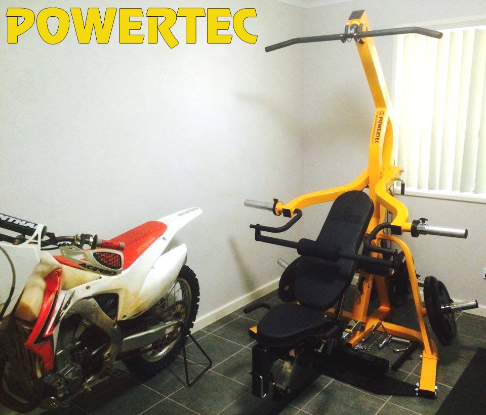 A compact room with a Powertec weight machine and a white and red dirt bike parked next to it. The weight machine is bright yellow with black padding and adjustable weights. The room has tiled flooring and a window with vertical blinds.