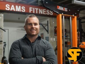 A person standing in a gym with arms crossed, smiling. Theres a weight machine in the background and a sign reading SAMS FITNESS. A logo with the letters SF is visible in the corner.