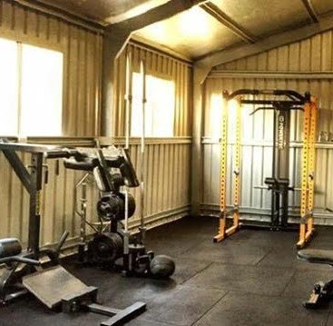 A home gym with various equipment, including a squat rack, weight bench, and a stack of weights. The room has metal walls and overhead lights, creating a functional workout space.