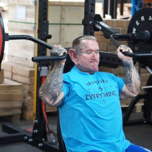 A person with tattoos is seated in a gym, lifting weights on a shoulder press machine. They wear a blue shirt with the text Mindset is Everything. Cardboard boxes are stacked in the background.