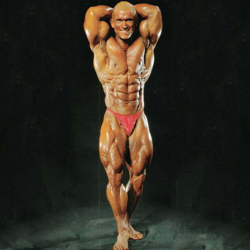 A bodybuilder posing on a dark background, wearing red trunks. The individual has a muscular build, showcasing defined abs and biceps, with hands clasped behind the head and legs slightly apart, highlighting their effort to build massive legs.