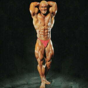 A bodybuilder posing on a dark background, wearing red trunks. The individual has a muscular build, showcasing defined abs and biceps, with hands clasped behind the head and legs slightly apart, highlighting their effort to build massive legs.