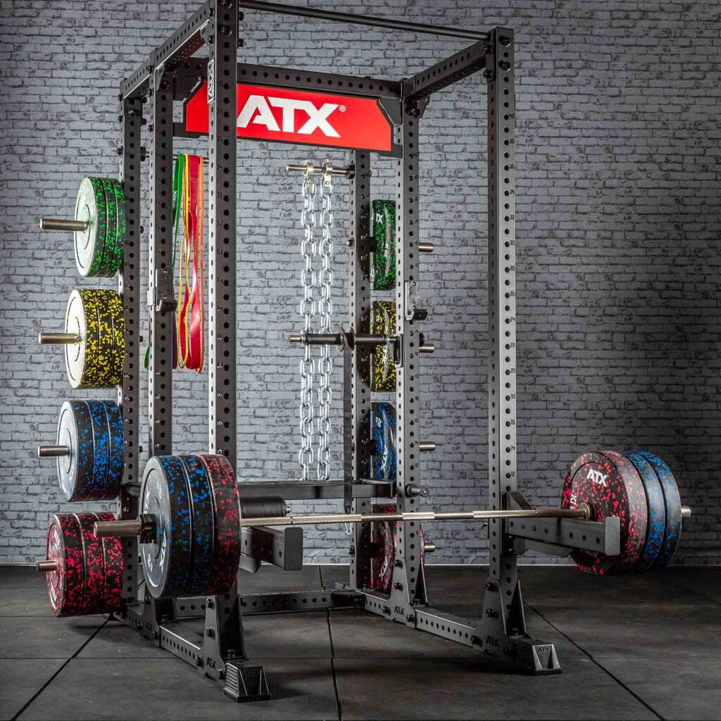A gym setup featuring a power rack with multiple weight plates and chains. The equipment is positioned against a brick wall, with a red ATX logo above the rack. The weights have a colorful speckled design.