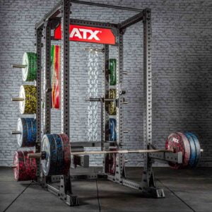 A gym setup featuring a power rack with multiple weight plates and chains. The equipment is positioned against a brick wall, with a red ATX logo above the rack. The weights have a colorful speckled design.