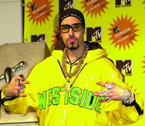 A person wearing a shiny yellow jacket with WESTSIDE printed in green, large sunglasses, and multiple rings and chains poses in front of a backdrop featuring the MTV Europe Music Awards 2001 logo.