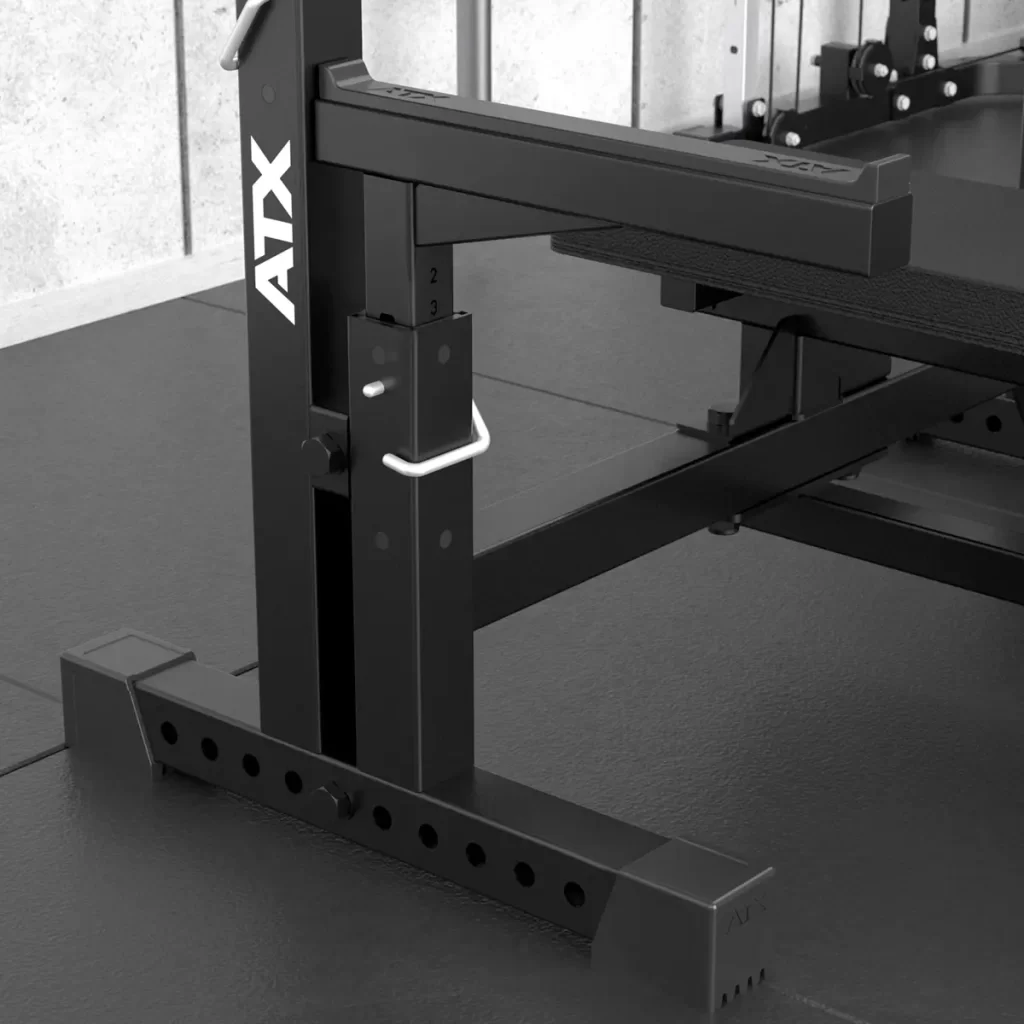 Close-up of the ATX® Olympic Bench Press 700 frame, showcasing its adjustable mechanism with multiple settings. The equipment is placed on a dark, textured floor in a fitness area.