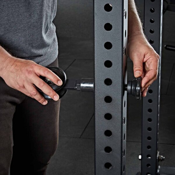 A person is adjusting the safety spotter arms on a metal rack in a gym, tightening the knobs to ensure secure placement.