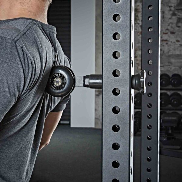 A person uses the ATX® Mobility Roller attached to a gym rack for their upper back. They wear a gray shirt, and exercise equipment and weights are visible in the background.