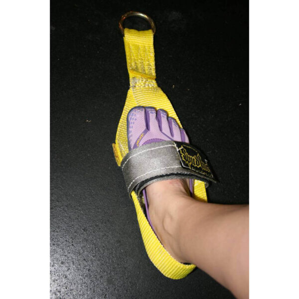 A foot wearing a purple and gray Vibram five-finger shoe is inserted into a yellow fabric harness with a metal ring at the top. The harness is lying flat on a black surface.