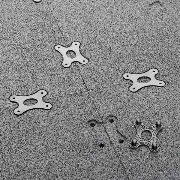 Silver-metal, four-point star brackets from the GymFloor RTS 50x50@20mm System are scattered on a textured gray surface. Nearby, shadows of two small black screws add contrast.