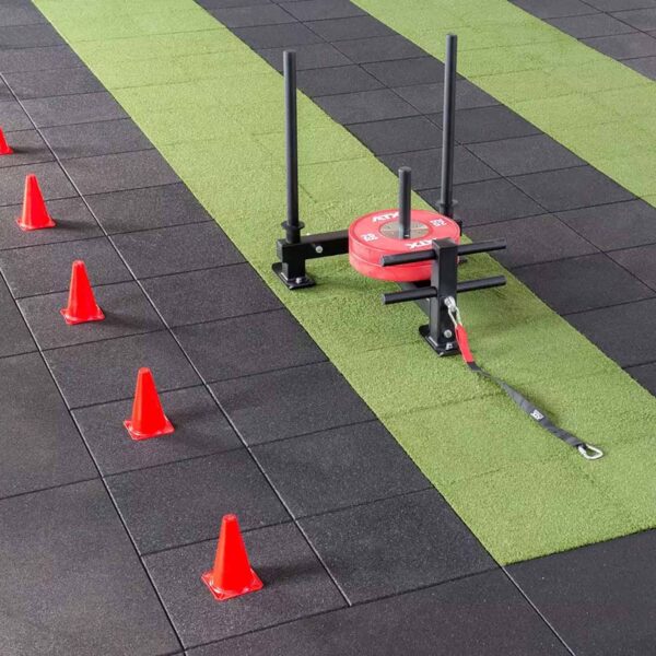 A gym sled with red weights and a harness rests on a green-and-black GymFloor RTS 50x50@20mm System. Red cones are aligned beside the sled, indicating a training path.