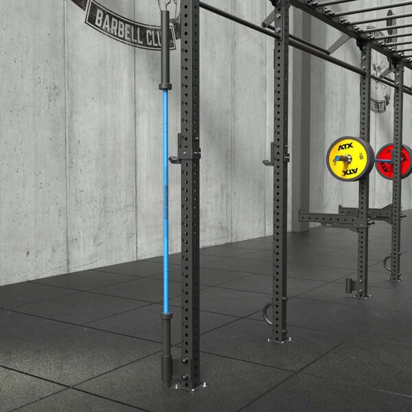 A gym setup with a gray concrete wall features squat racks sporting yellow ATX and red XV plates, a barbell storage offset, and a blue band on one rack. The floor is covered in black mats.