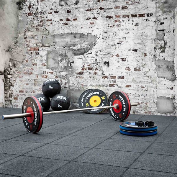 In a gym setting, a barbell loaded with weight plates rests on the Shock & Sound Tile 30mm System rubber mat. Additional weight plates and medicine balls are neatly arranged against a backdrop of a brick wall with peeling paint.