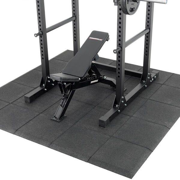 A black adjustable workout bench set on Shock & Sound Tile 30mm System beneath a power rack, designed for weightlifting exercises with barbell supports and safety pins.
