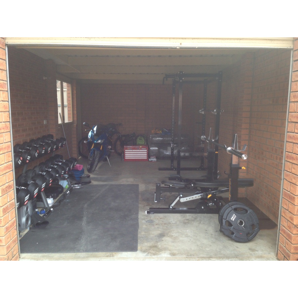 The image shows a garage partially open, revealing a home gym setup with a squat rack, weight plates, and dumbbells on the left. On the right, a motorcycle is parked near a red toolbox against the brick walls.
