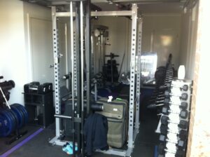 Lee Priests garage gym is a home fitness haven, boasting an array of workout equipment: weight plates, dumbbells, a full rack, exercise machines, and even a suitcase. A jacket is draped over the rack in this compact space where fitness items are impeccably organized.