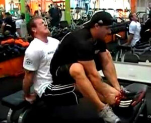 Two men are at a gym. One man is seated on a bench performing tricep dips while the other man sits on his lap, providing extra resistance. They are surrounded by gym equipment and other people working out.