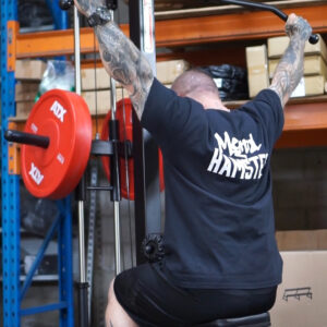 A tattooed individual is using a lat pulldown machine in a gym. They are wearing a black Mental Hamster t-shirt. Red weights are visible on the machine, and shelves with boxes can be seen in the background.