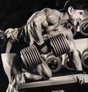 A muscular person, wearing a cap and shorts, lifts a heavy dumbbell while kneeling on a bench in a gym, surrounded by various weights.