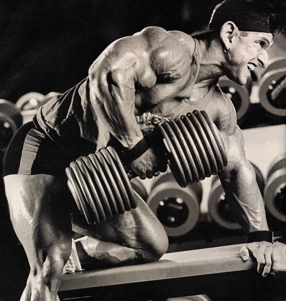 A muscular person, wearing a cap and shorts, lifts a heavy dumbbell while kneeling on a bench in a gym, surrounded by various weights.