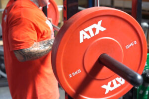 A person in a red shirt stands next to a barbell loaded with red ATX weight plates, each labeled 25 KG, in a gym setting. The background includes gym equipment and part of the persons tattooed arm is visible.