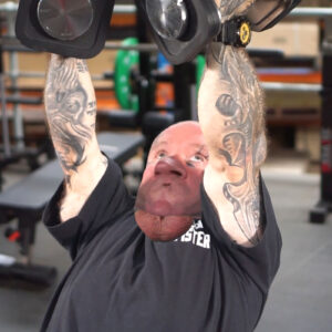 A person with tattooed arms is lying on a bench in a gym, performing dumbbell pec flys. The image is distorted, with the persons head and chin appearing unnaturally enlarged. Gym equipment can be seen in the background.