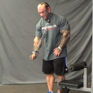 A muscular man in a gym performs a tricep workout using cable pulleys. He wears a gray t-shirt, black shorts, and blue sneakers. The background features a gray curtain and gym equipment.