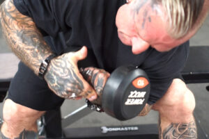 A heavily tattooed person performing a concentration curl with a 10 kg dumbbell, wearing a black shirt. The background features gym equipment.