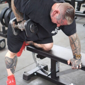 A heavily tattooed person with bleached hair and red sneakers is performing a one-arm dumbbell row in a gym. They kneel on a bench and focus intently on the exercise amidst gym equipment.