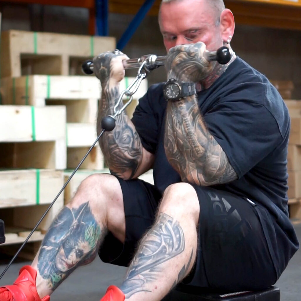 A heavily tattooed man is seated and performing a bicep workout using a cable machine in a gym. He is wearing a black T-shirt, black shorts, and red shoes. Wooden crates are visible in the background.