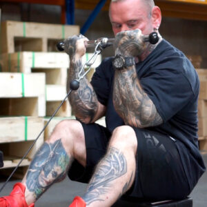 A heavily tattooed man is seated and performing a bicep workout using a cable machine in a gym. He is wearing a black T-shirt, black shorts, and red shoes. Wooden crates are visible in the background.