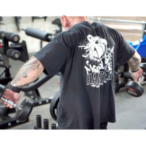 A person with tattoos is lifting dumbbells in a gym. They are wearing a black t-shirt with a graphic design on the back. Gym equipment is visible in the background.