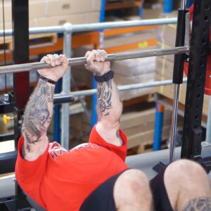 A person in a red shirt with tattooed arms is performing a bench press in a gym. They are lying on a bench, gripping the barbell above them, with equipment and stacked boxes visible in the background.