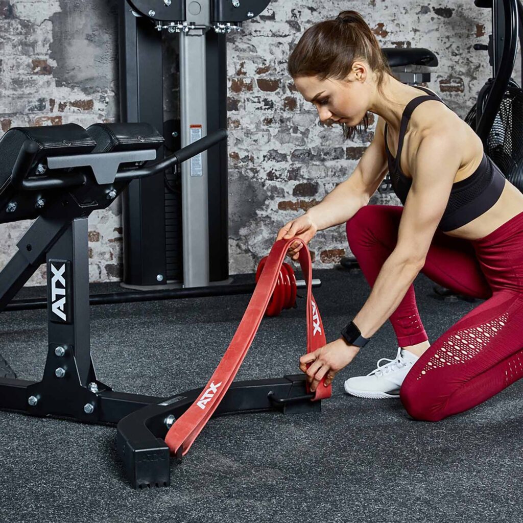 A woman in workout attire kneels on the gym floor, attaching a red resistance band to the ATX® Hyperextension Machine 650. She is focused on setting it up, with various gym machinery and a brick wall in the background.