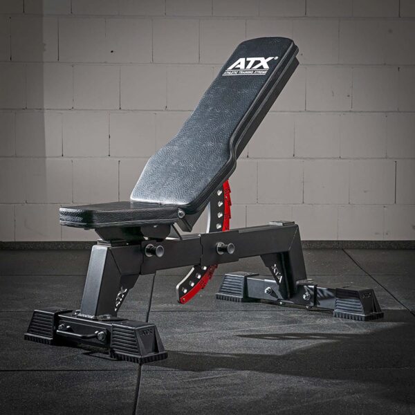 An ATX® Utility Bulls Bench, adjustable and black with a red adjustment bar, sits partially inclined on a gym floor against a white brick wall.