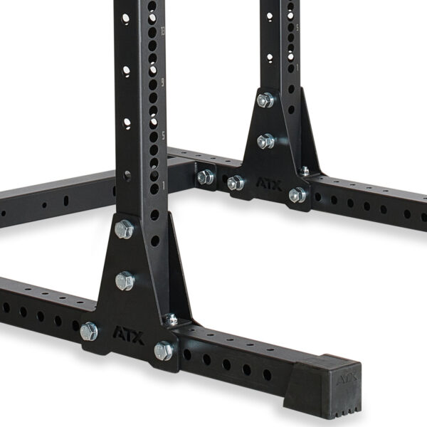 A close-up of the ATX® Power Rack 755 Short Distance shows a black metal stand with multiple holes and bolts. It features a sturdy base with a rectangular foot and rubber caps for stability, designed to support heavy equipment.